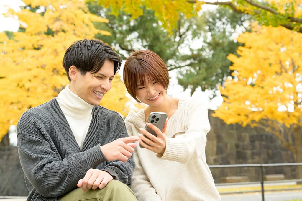 紅葉の中で仲良くスマホを眺める男女。マッチングアプリの追いメッセージが成功し、良好な関係を築けたイメージ画像。