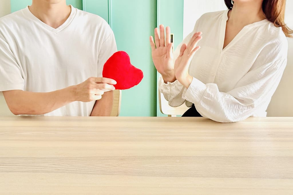 男性が差し出した赤いハートを、女性が両手で遮って拒否している様子。マッチングアプリでの失恋やお断りサインを表現した画像。