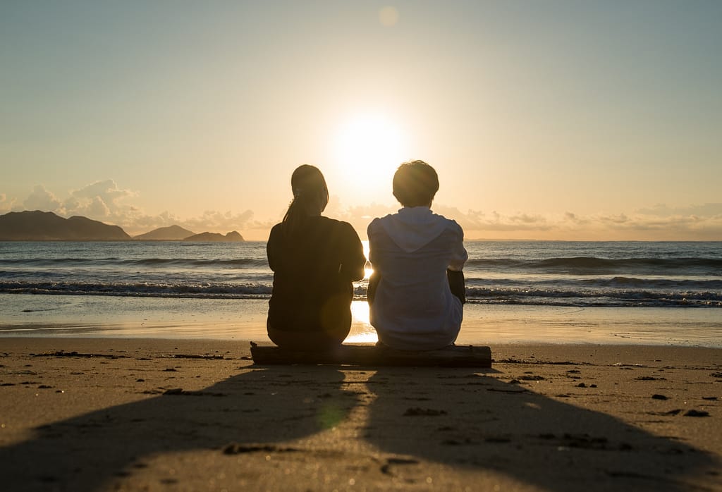 マッチングアプリで理想の恋人と出会い、夕日の砂浜で仲良く座っているカップルの後ろ姿。